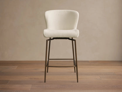 Kirsten Swivel Stool with cream upholstered curved back and seat, tall dark bronze iron legs, and footrest on a wooden floor.