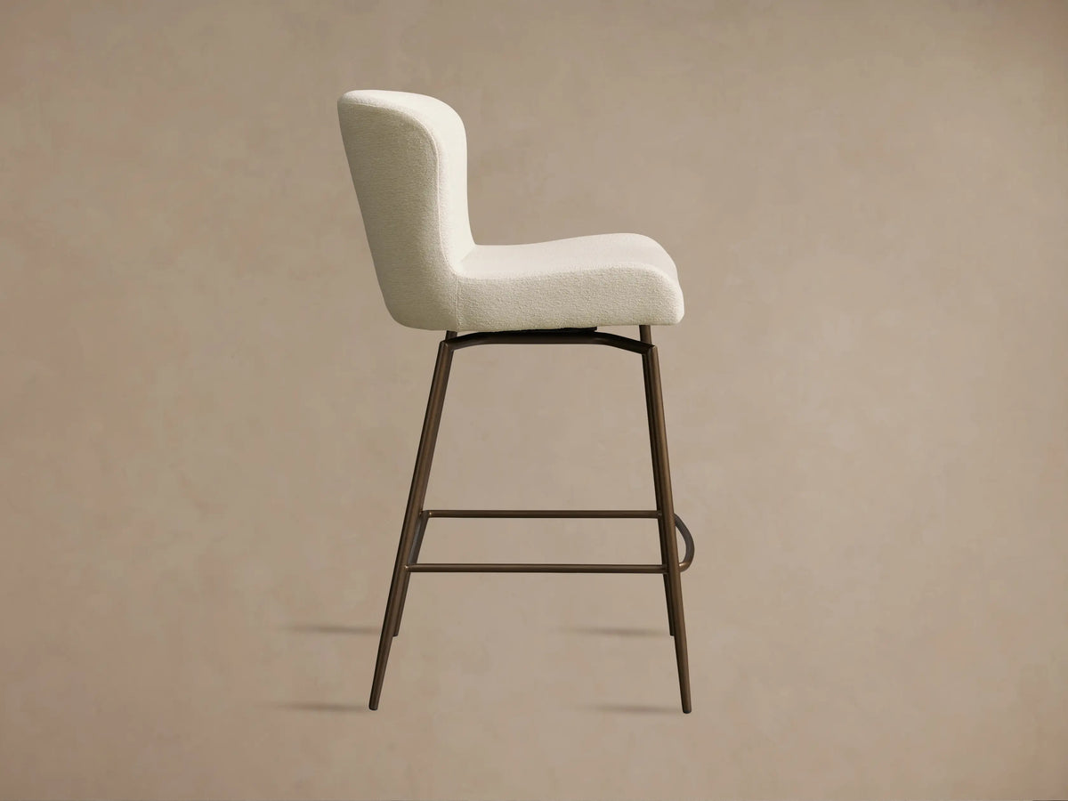 Kirsten Swivel Stool with a cream upholstered seat and backrest, iron legs in a bronze finish, shown in a side angle view.