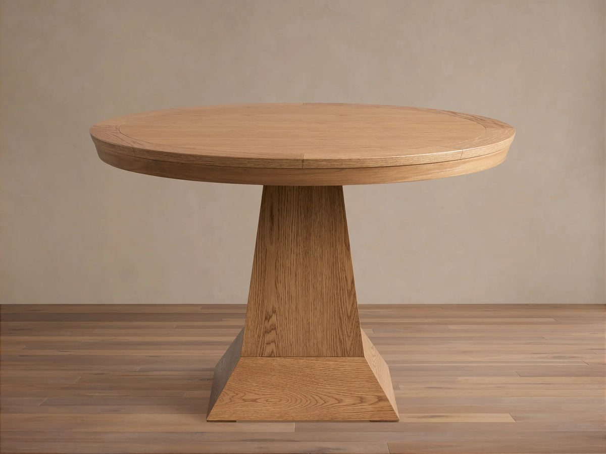 Leighton Round Dining Table in oak finish, featuring a smooth circular top and a wide, tapered pedestal base on a wood floor.