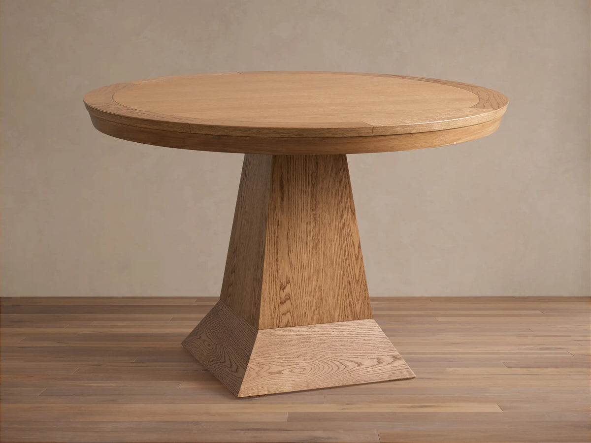 Leighton Round Dining Table in oak finish, featuring a smooth circular top and a wide, tapered pedestal base on a wood floor.