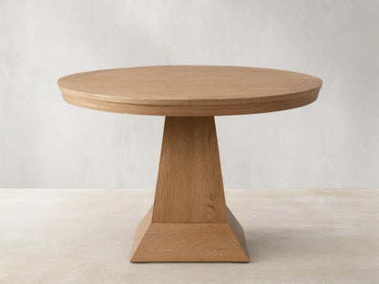 Leighton Round Dining Table in oak finish, featuring a smooth circular top and a wide, tapered pedestal base on a wood floor.