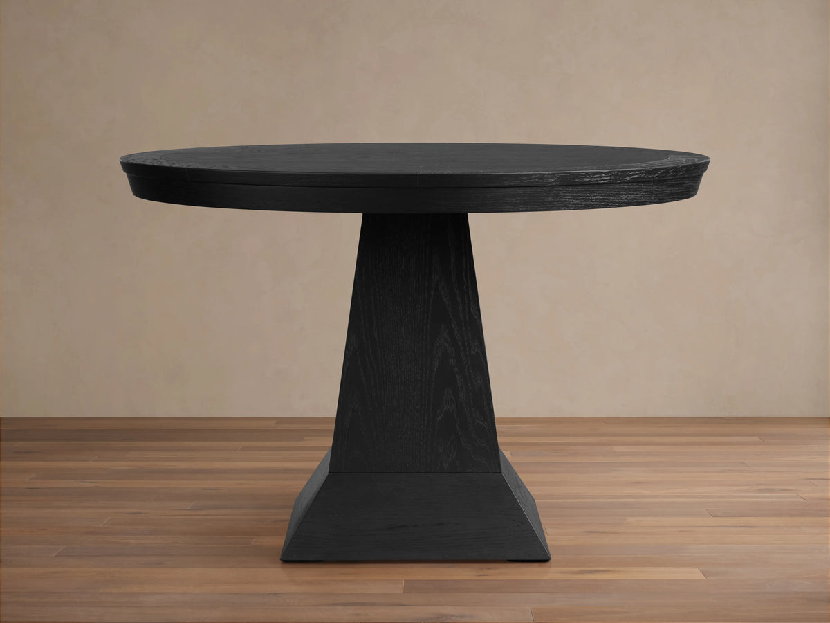 Leighton Round Dining Table in dark wood finish on a pedestal base. The table is placed on a wooden floor with a beige background.