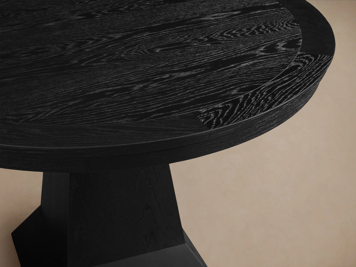 Leighton Round Dining Table in dark finish with a smooth, circular top and a sturdy, tapered pedestal base, displayed on wooden flooring.