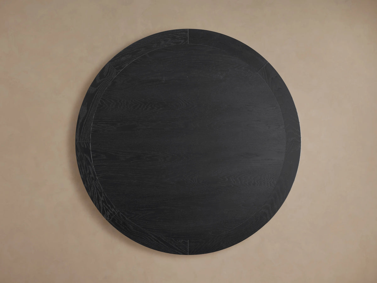 Leighton Round Dining Table with dark oak veneer finish, showcasing wood grain patterns. Supported by a geometric pedestal base.