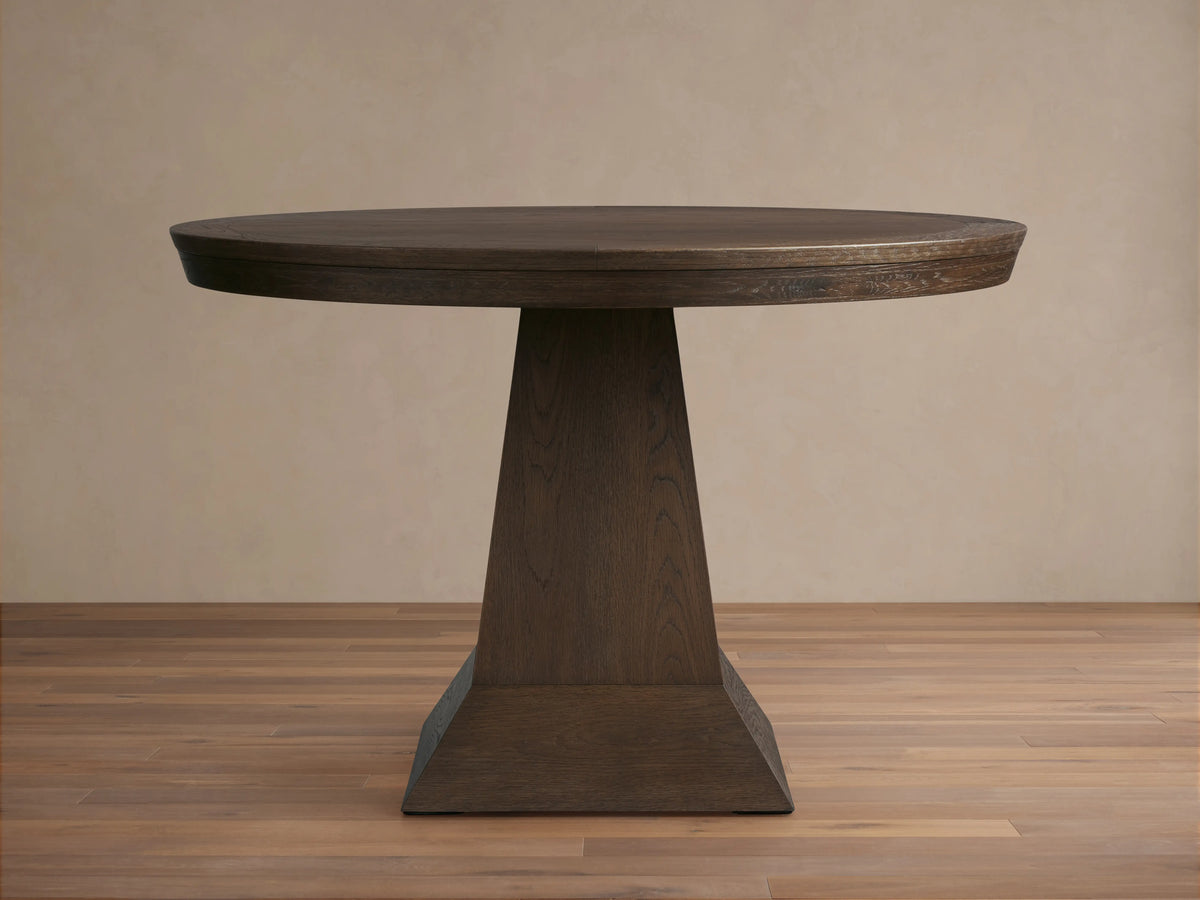 Leighton Round Dining Table made of dark oak veneers with a smooth circular top and a broad, geometrically tapered base, displayed on hardwood flooring.