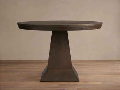 Leighton Round Dining Table made of dark oak veneers with a smooth circular top and a broad, geometrically tapered base, displayed on hardwood flooring.