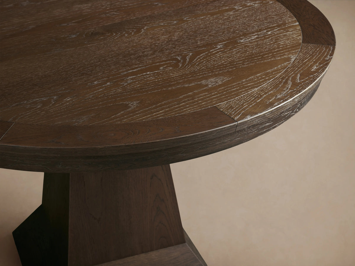 Round wooden dining table with dark brown oak veneer finish, featuring a thick pedestal base. Positioned on a wooden floor against a beige wall.