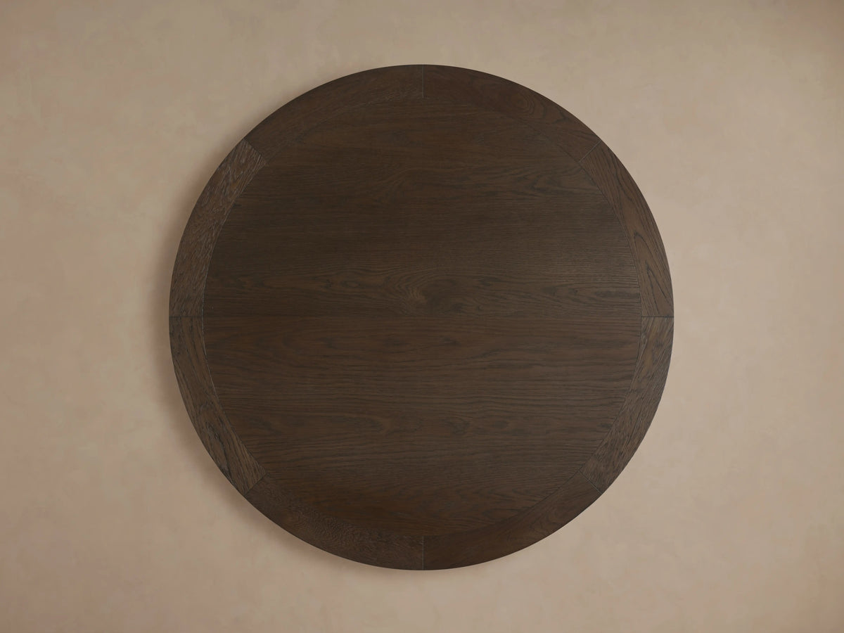Round dining table with dark oak veneer and visible wood grain, featuring a thick edge and sturdy pedestal base on a neutral background.