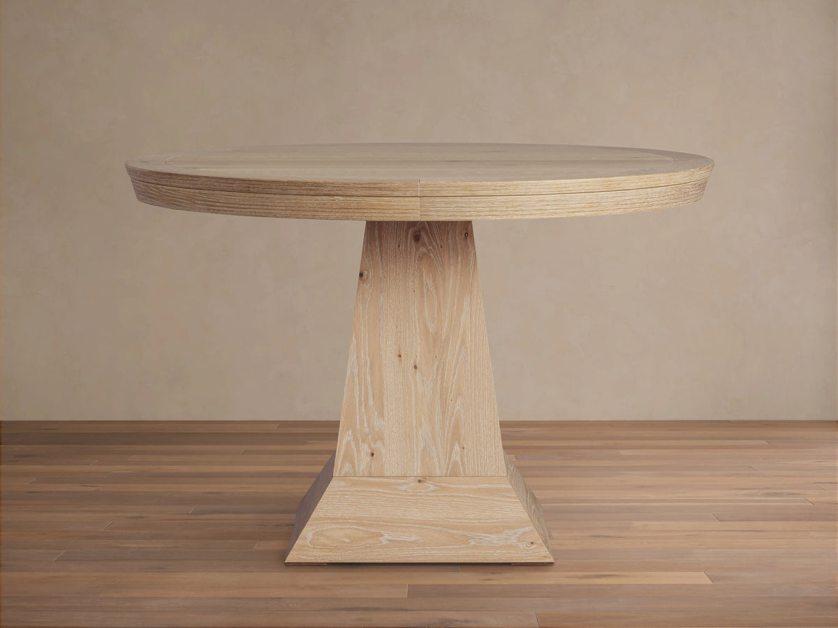 Leighton Round Dining Table with a circular oak veneer top and a unique tapered pedestal base, set on a wooden floor.