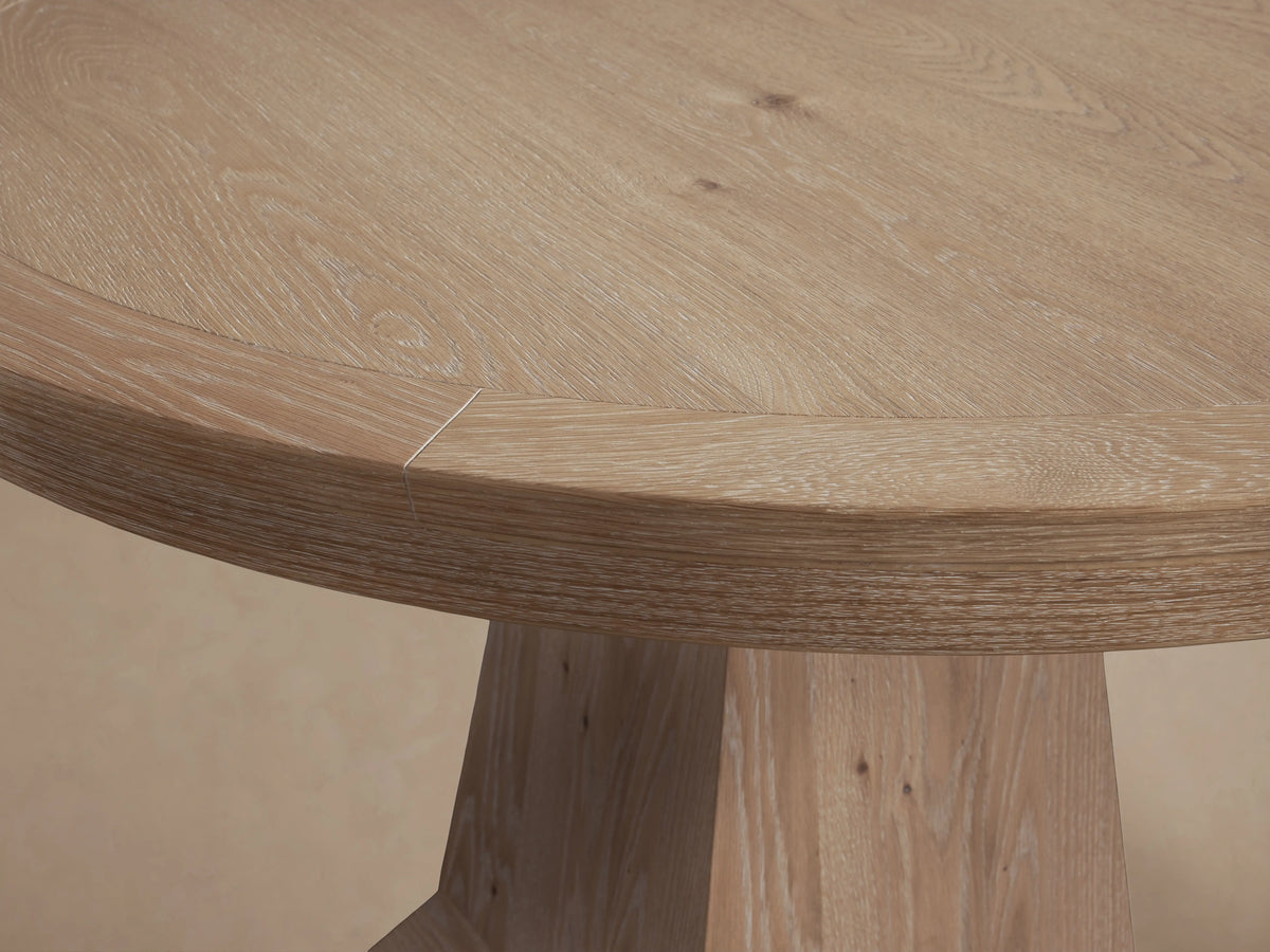 Leighton Round Dining Table with oak veneer, round top, and wide, tapered pedestal base on hardwood floor. Light wood finish.