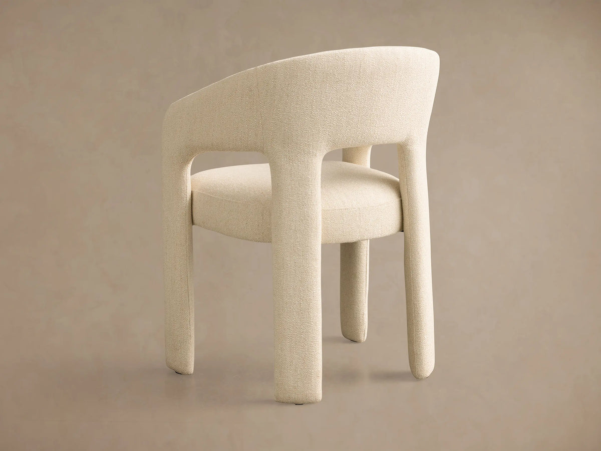 Side profile of a cream Lilette Dining Arm Chair, fully upholstered in textured fabric with a curved back, armrests, and thick legs.