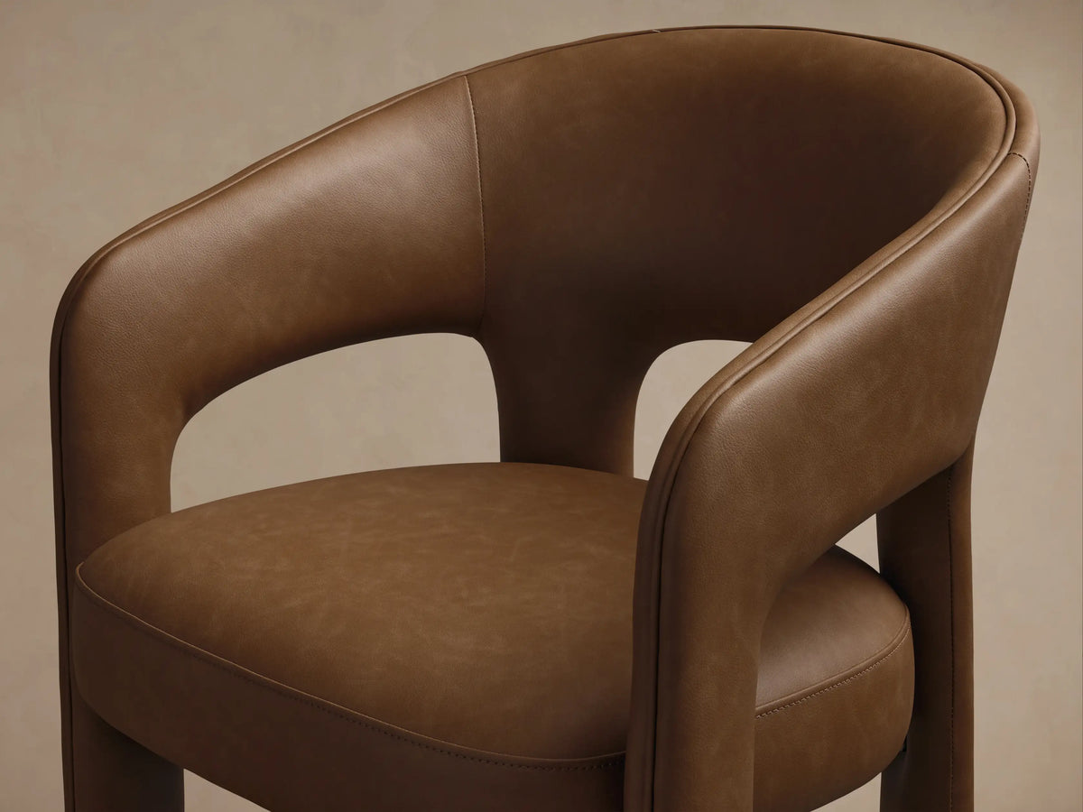 Rear three-quarter view of brown Lilette Faux Leather Dining Arm Chair, with curved backrest, integrated armrests, thick legs.