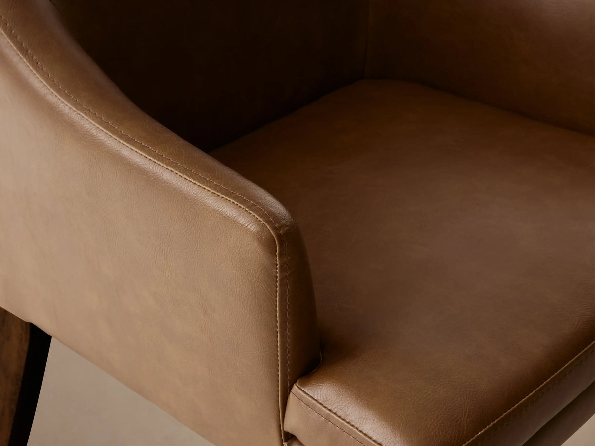Brown faux leather dining armchair with gently curved backrest and armrests, on wooden legs, set against a wooden floor and beige wall.