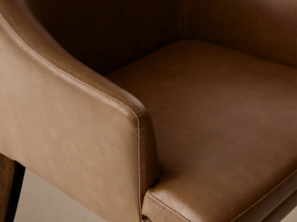 Brown faux leather dining armchair with gently curved backrest and armrests, on wooden legs, set against a wooden floor and beige wall.
