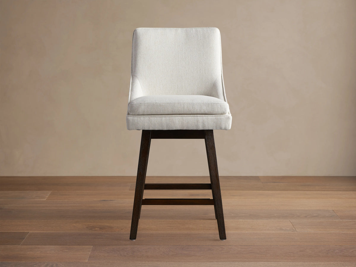 Lunden Swivel Stool with curved off-white upholstered seat and back, on a dark wood frame, angled right on a wood floor.