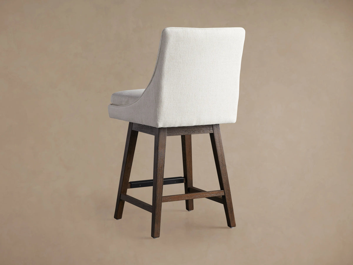 Lunden Swivel Stool, close-up of light beige textured fabric seat and elegantly curved backrest. A dark wooden leg is visible.