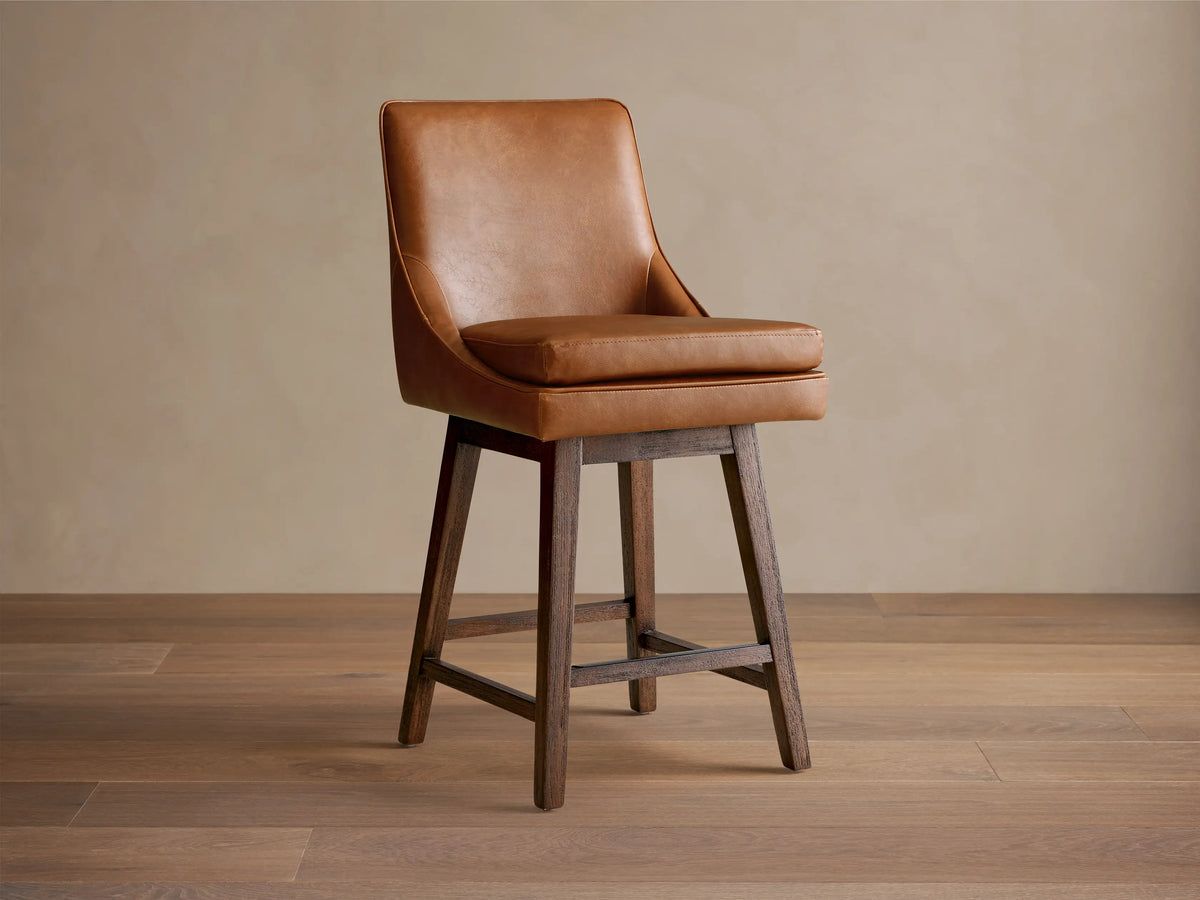 Lunden Faux Leather Swivel Stool in warm brown leather. Features a curved backrest, cushioned seat, and dark wooden legs on a hardwood floor.