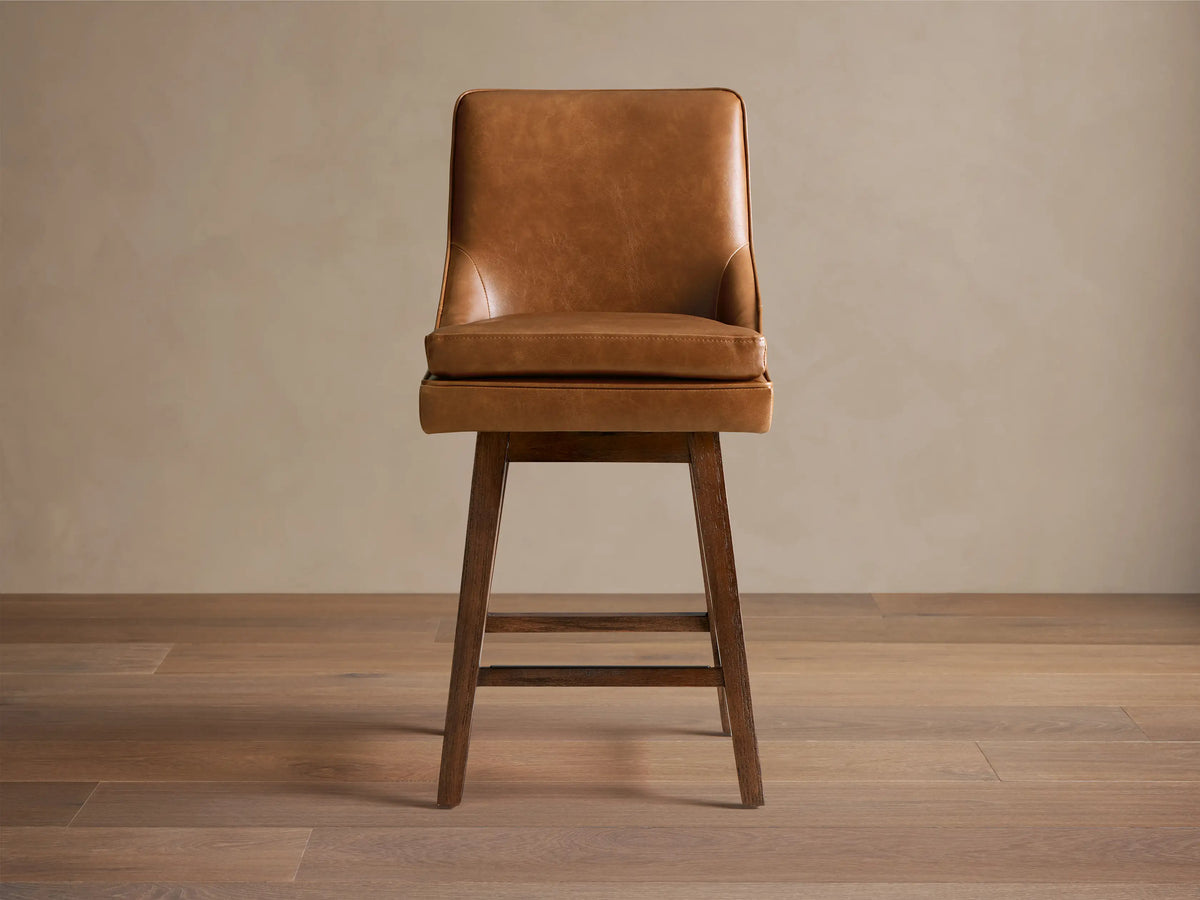 Lunden Faux Leather Swivel Stool in warm brown leather. Features a curved backrest, cushioned seat, and dark wooden legs on a hardwood floor.
