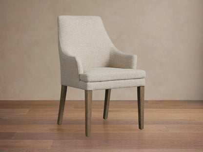 Lunden Dining Arm Chair in Fawn features a high, gently curved backrest, upholstered in a light beige fabric with dark wooden legs on a wooden floor.