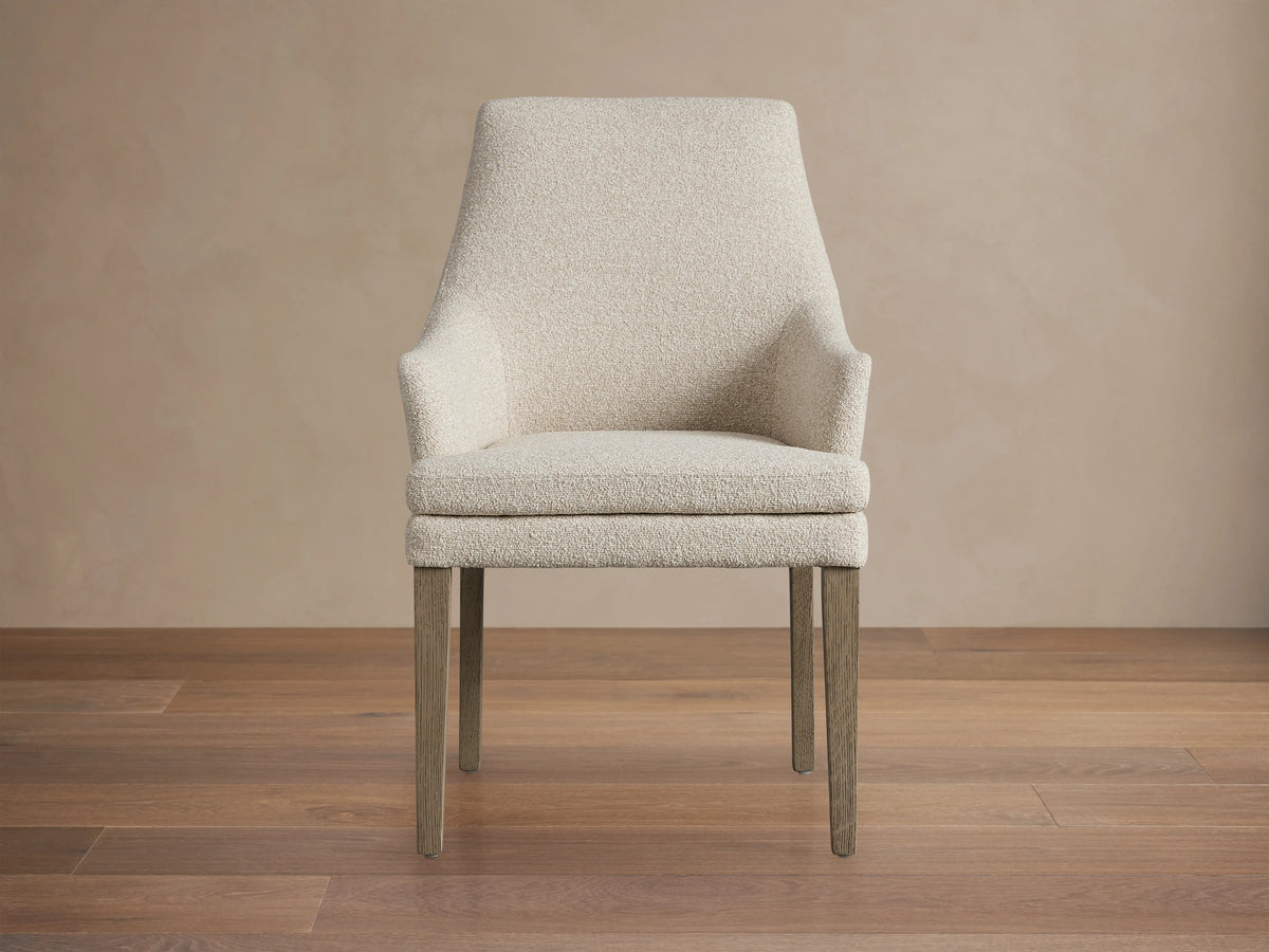 Lunden Dining Arm Chair in Fawn features a high, gently curved backrest, upholstered in a light beige fabric with dark wooden legs on a wooden floor.