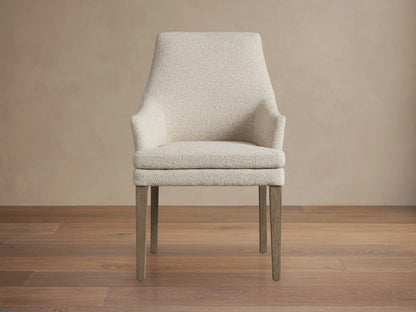 Lunden Dining Arm Chair in Fawn features a high, gently curved backrest, upholstered in a light beige fabric with dark wooden legs on a wooden floor.