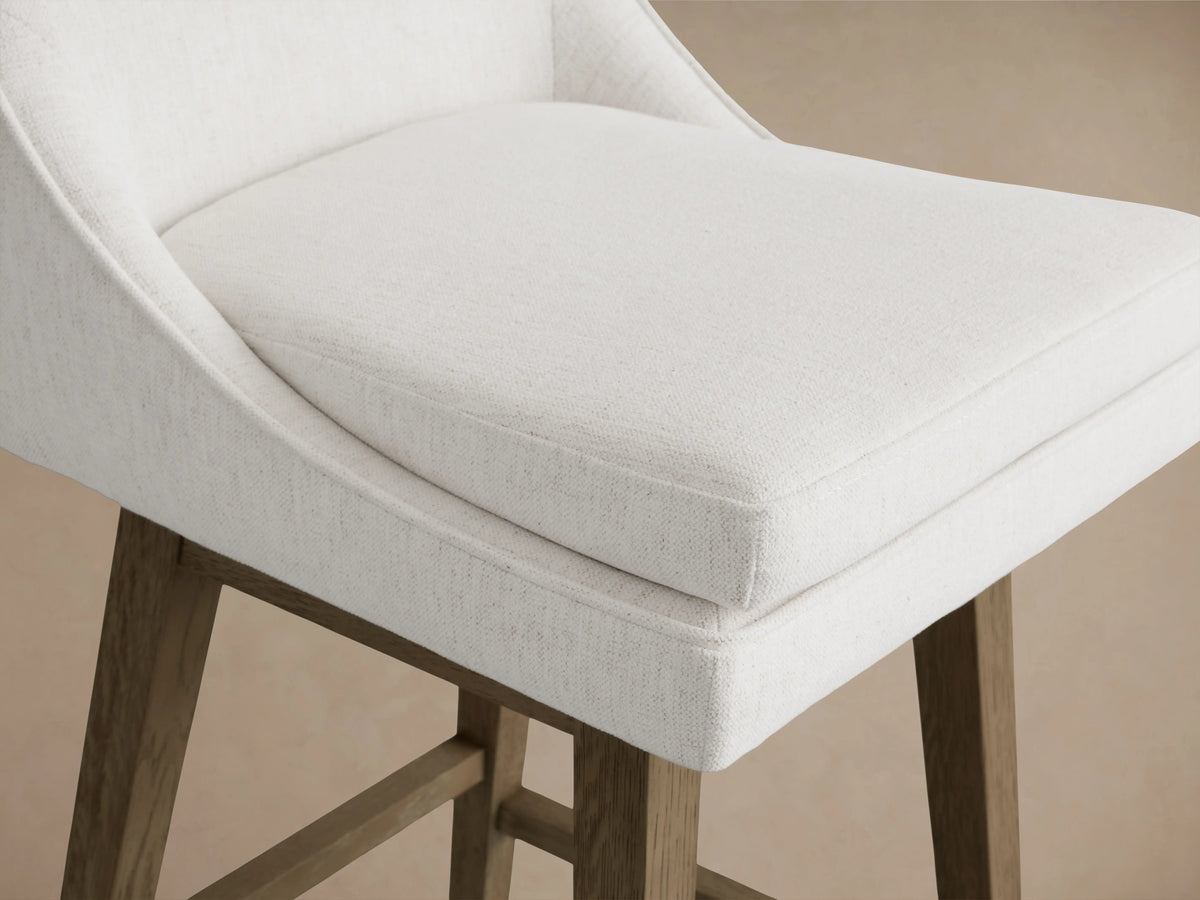 Lunden Swivel Stool in Fawn with a curved backrest, upholstered in light fabric, on a wooden base with footrest, shown on a wood floor.