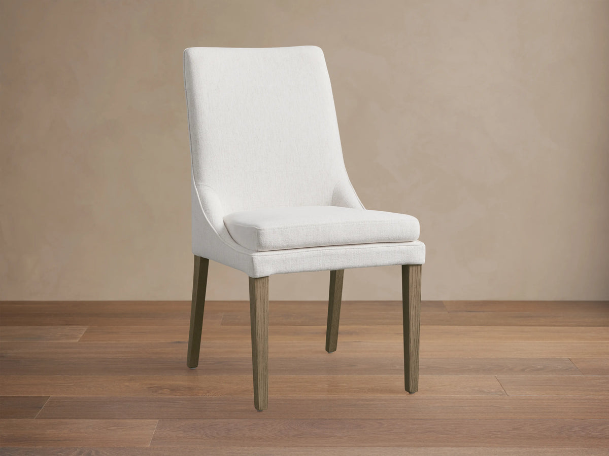 Lunden Dining Side Chair in Fawn with a curved backrest, upholstered in soft ecru fabric, and wooden legs on a hardwood floor.