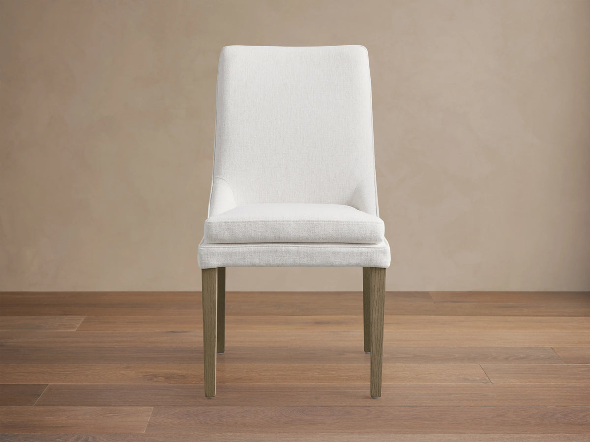 Lunden Dining Side Chair in Fawn with a curved backrest, upholstered in soft ecru fabric, and wooden legs on a hardwood floor.