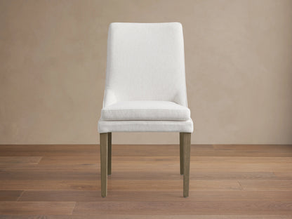 Lunden Dining Side Chair in Fawn with a curved backrest, upholstered in soft ecru fabric, and wooden legs on a hardwood floor.