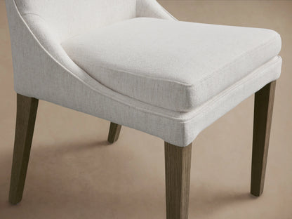 Lunden Dining Side Chair in Fawn shown from the front, features a softly curved backrest, cushioned seat, and wooden legs on a wooden floor.