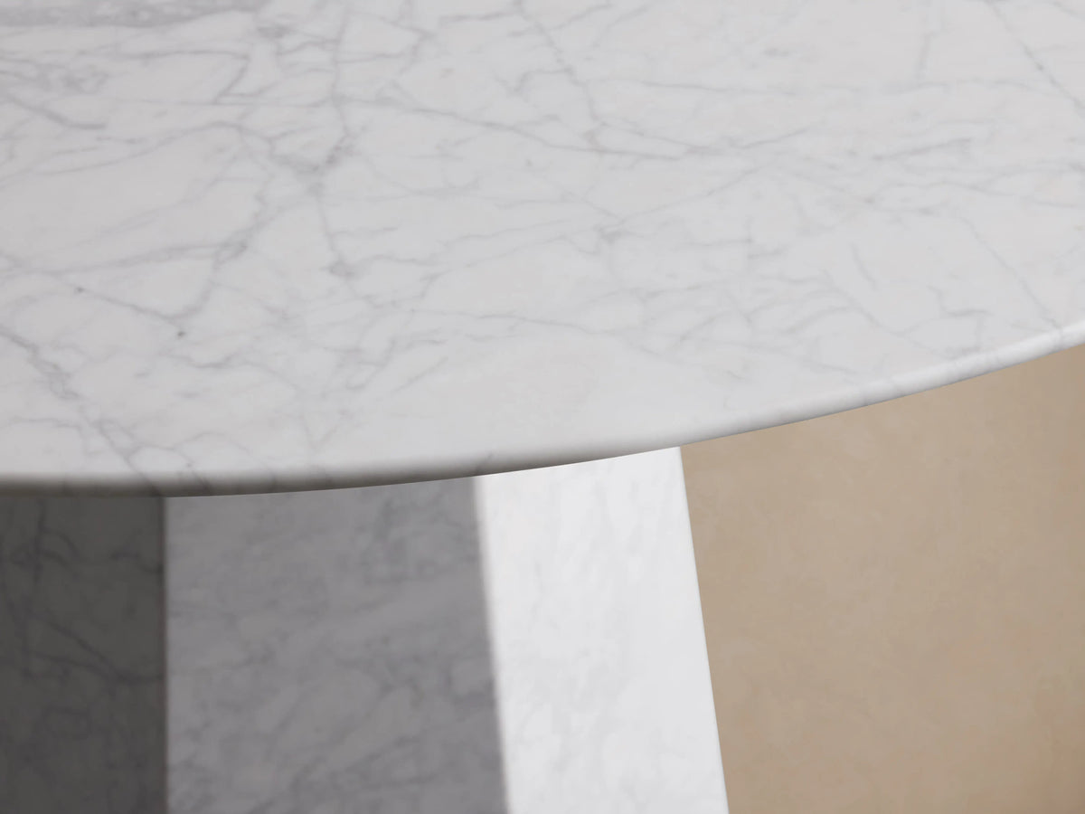Modena Dining Table. Round white Carrara marble top on a geometric tapered pedestal base, on a wooden floor against a beige wall.