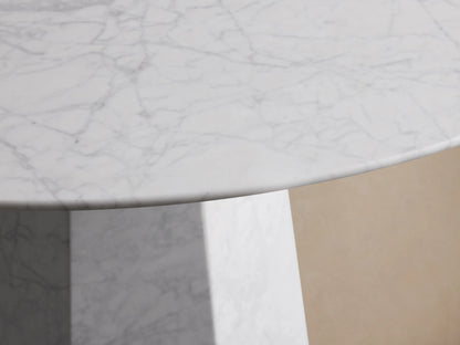 Modena Dining Table. Round white Carrara marble top on a geometric tapered pedestal base, on a wooden floor against a beige wall.