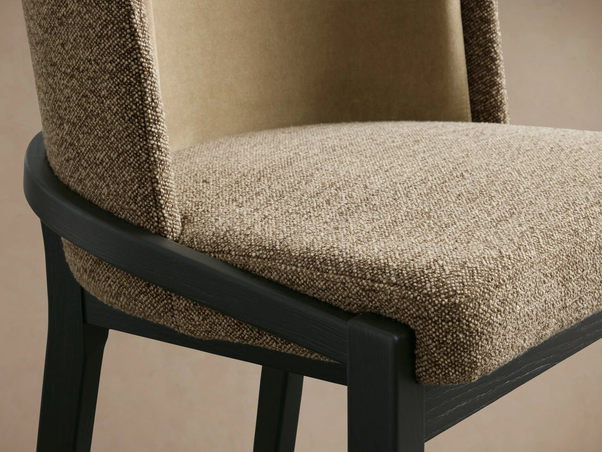 The Mena Stool features a textured beige fabric with a dark wood frame. The curved backrest and seat display soft, plush upholstery.
