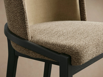 The Mena Stool features a textured beige fabric with a dark wood frame. The curved backrest and seat display soft, plush upholstery.
