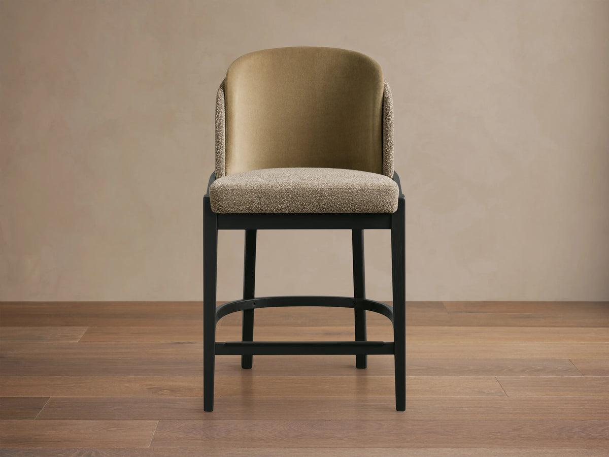 Beige faux leather and taupe fabric Mena Stool with Vesuvio Black ash wood frame, on a wooden floor, against a neutral wall.