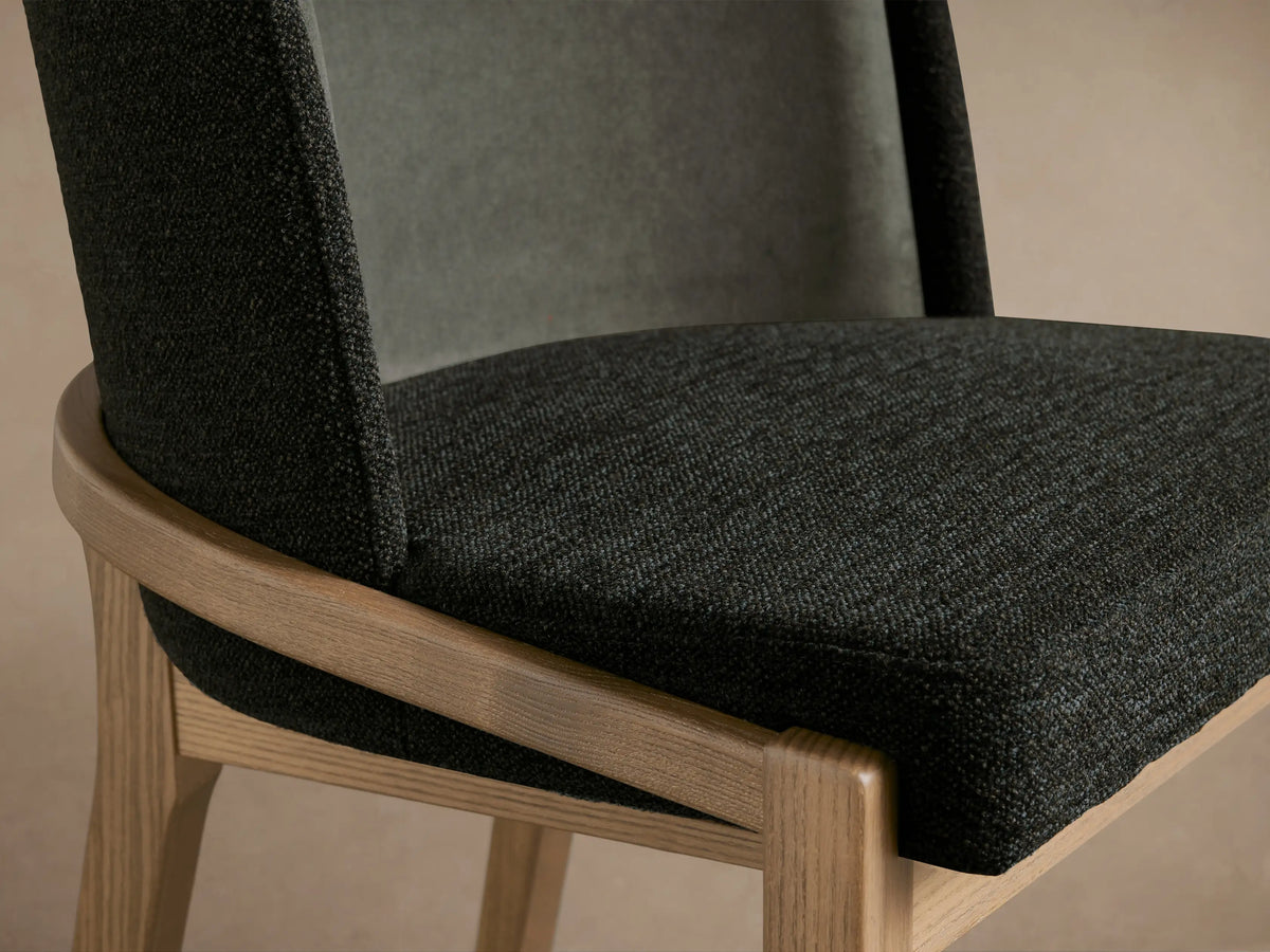 Close-up, angled view of Mena Dining Chair with dark grey textured fabric seat and layered back, light ash wood frame.