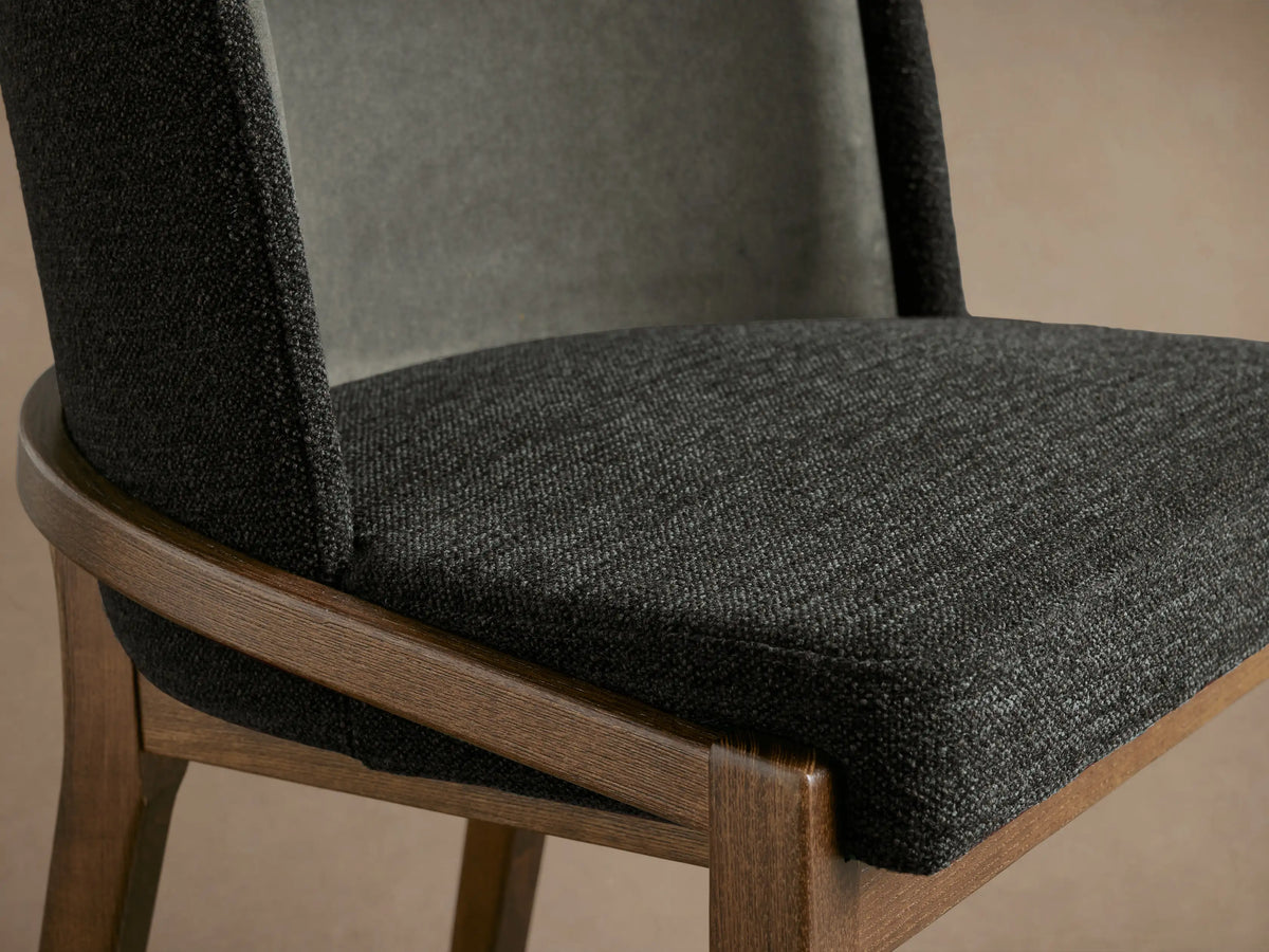 Angled view of Mena Dining Chair: dark gray fabric seat, lighter gray inner back, brown ash wood frame, tapered legs.