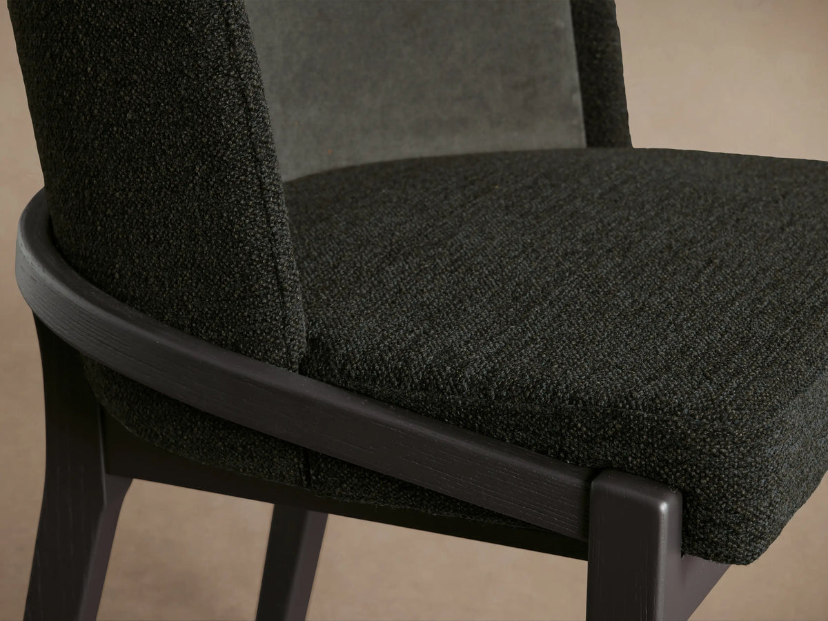 Close-up, angled view of Mena Dining Chair. Dark gray textured fabric seat, dark ash wood frame, layered backrest in two fabrics.