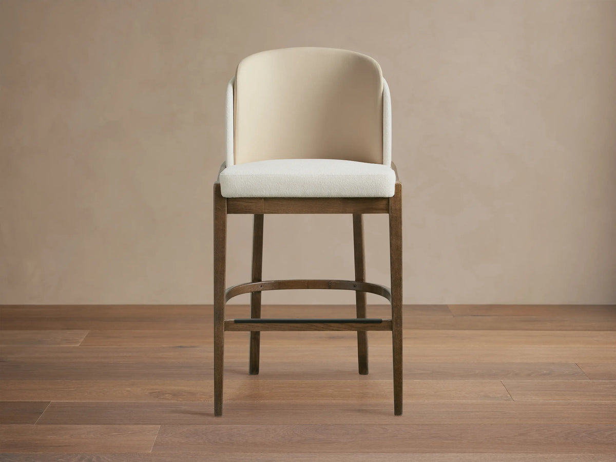 Mena Stool: Dark ash wood frame, cream upholstered seat, two-tone layered curved back, tapered legs. On wooden floor.