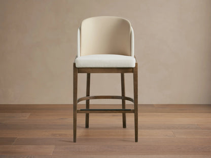 Mena Stool: Dark ash wood frame, cream upholstered seat, two-tone layered curved back, tapered legs. On wooden floor.