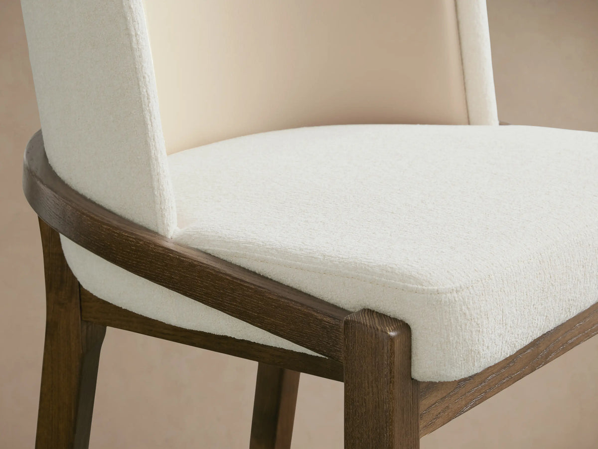 Mena Stool, front view. Dark wooden frame, light cream upholstered seat. Curved backrest with cream inner & lighter outer fabric, on wood floor.