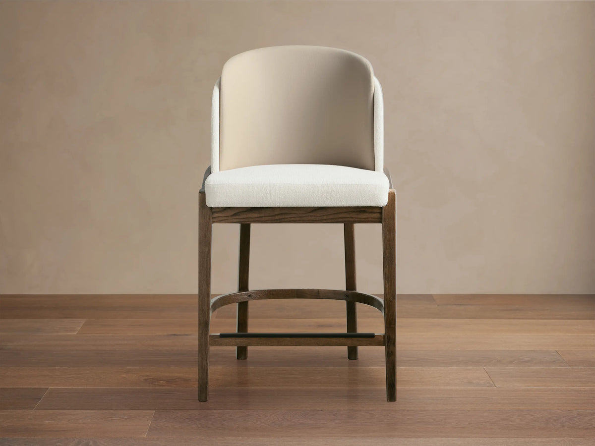 Mena Stool, dark ash wood frame with tapered legs, light cream upholstered seat & two-tone beige backrest, on wood floor.