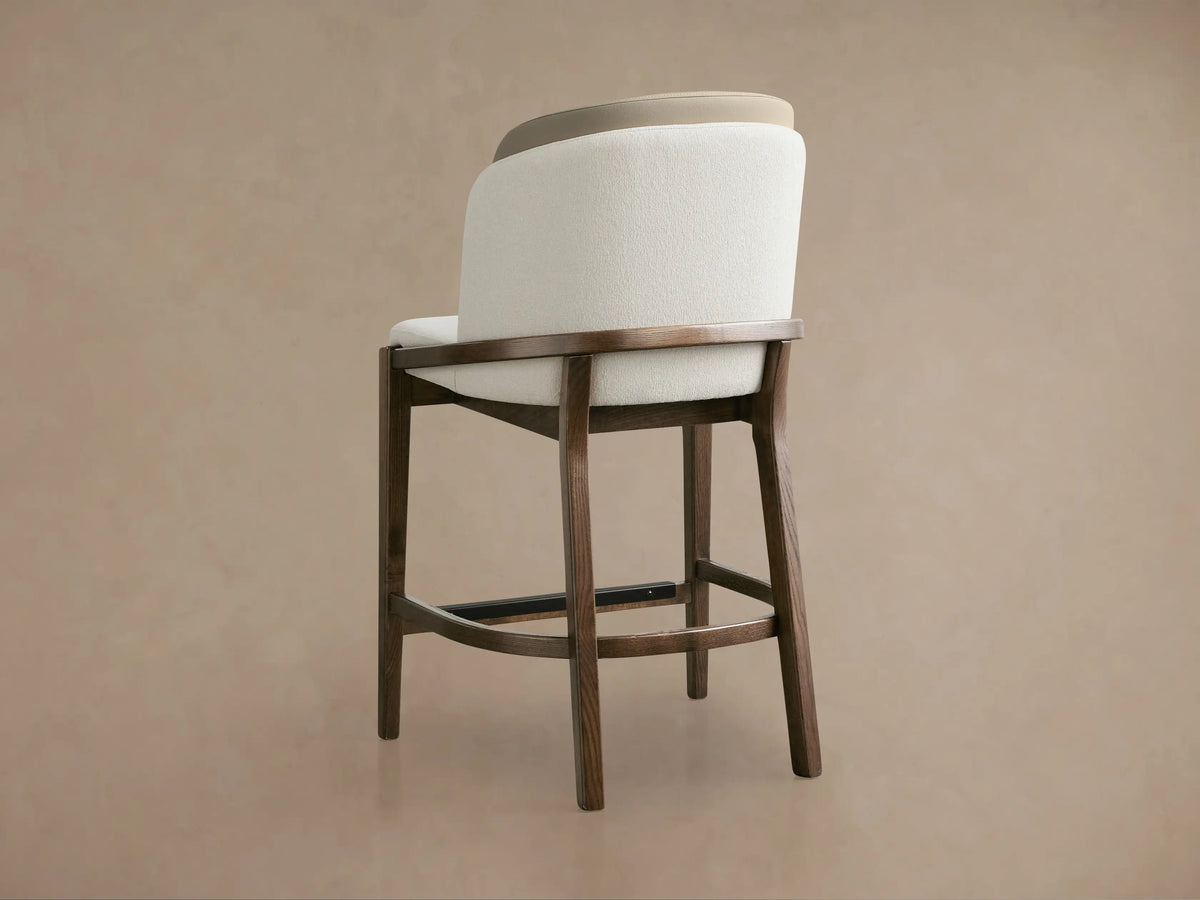 Close-up of Mena Stool with textured cream upholstered seat & back, smooth tan inner back, and elegant dark ash wood frame.