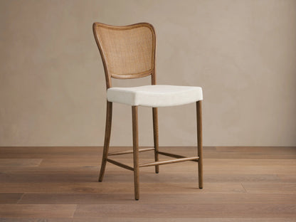 Noa Stool, cinder-stained oak frame, woven rattan back, light cream fabric cushion, angled right on wood floor.