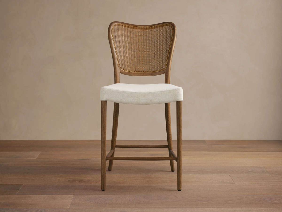 Noa Stool, cinder-stained oak frame, woven rattan back, light cream fabric cushion, angled right on wood floor.