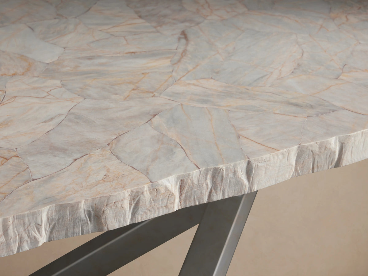 Petra Dining Table with a buffed, polished stone tabletop atop an X-shaped, iron base in dark grey, on a wooden floor.