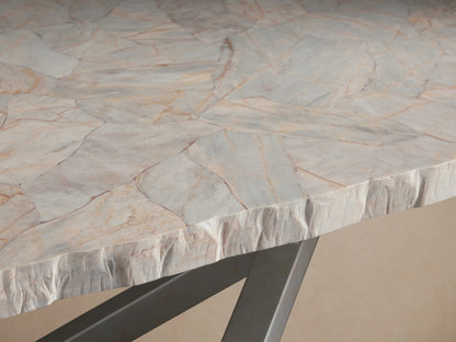 Petra Dining Table with a buffed, polished stone tabletop atop an X-shaped, iron base in dark grey, on a wooden floor.