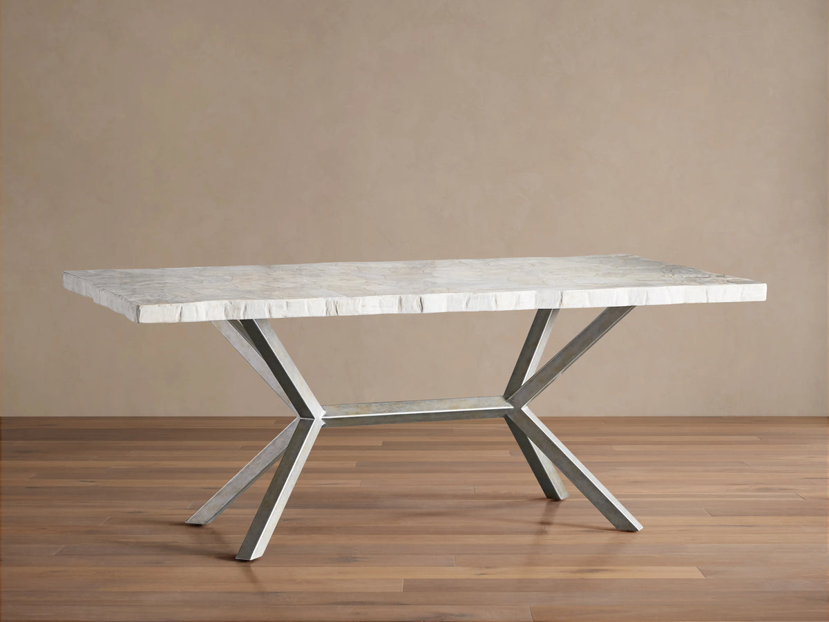 Rectangular Petra Dining Table with a textured grey stone top and crossed iron legs, featuring an antiqued dark grey patina, on a wooden floor.