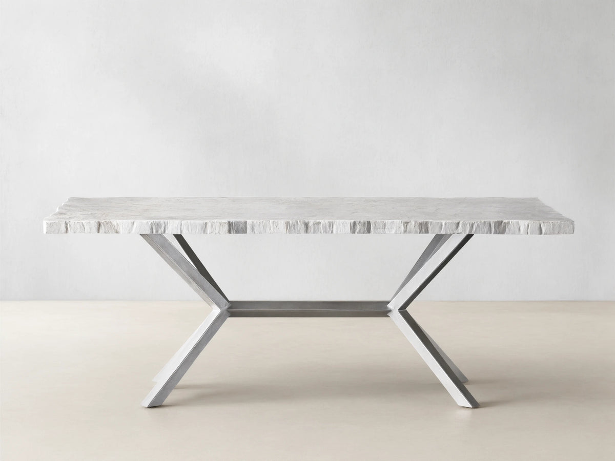 Rectangular Petra Dining Table with a textured grey stone top and crossed iron legs, featuring an antiqued dark grey patina, on a wooden floor.