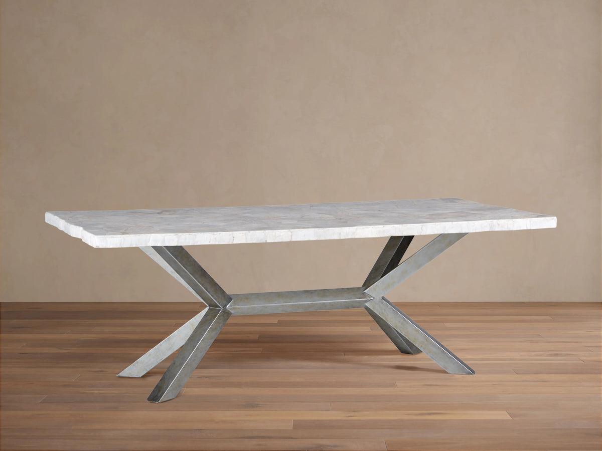 Petra Dining Table with a polished, light stone top and iron base in an X-shape, featuring a dark grey patina, placed on wooden flooring.